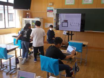 一人の男の子がタブレットを触り、斜め前の机には生徒2名が集まっている教室内での写真