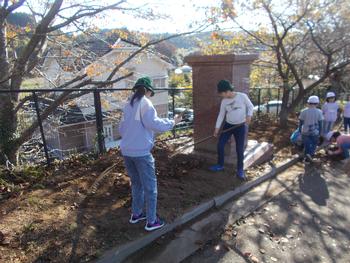 レーキを使いながら落ち葉を集めている高学年2名と奥で草取りをしている低学年の子供たちの写真