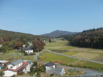 少し高台から写した、手前にある住宅の前に田畑が広がり、左右に少し赤く色づいた木が見える森、奥に山がある秋の風景を写した写真