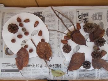 新聞紙の上に、丸い形の白い紙に落ち葉やドングリ、松ぼっくりで装飾がされ、目の部分がくりぬかれたお面が2つ並んでいる写真