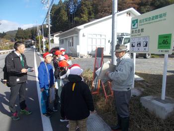 赤い帽子をかぶった小学生たちが先生と地域の人に災害時の避難場所について話を聞いている様子の写真