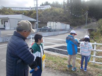 沢のフェンスの前で男の子がタブレットを使い、隣に女の子も立ちながら学習している様子を後方で見守る男性と生徒一人を写した写真