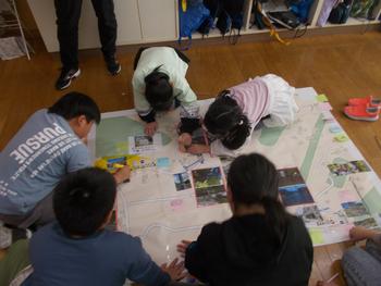 床に広げられた大きな地図の上に集まった数人の子どもたちが、写真や付箋を貼り付けたり、マーカーで印をつけたりしながら熱心に話し合っている様子の写真