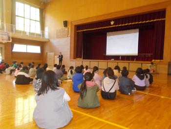 ステージ上の男性が子どもたちに向けてプレゼンテーションを行っており、子どもたちは床に座って話を聞いている様子の写真