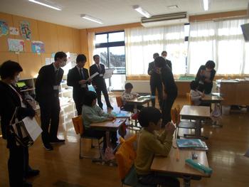 明るい教室で数名の小学生が個別に机に向かって学習しており、スーツ姿の大人たちが児童の様子を見学したり声をかけたりしている指導主事学校訪問の写真