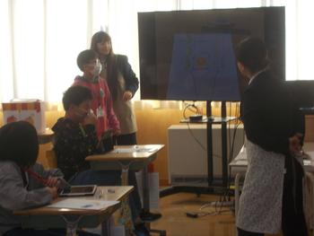 教室の前方で大きなモニターを使ってスライドを表示しながら、小学生が発表を行い、周囲の児童や教員が真剣に話を聞いている様子の写真