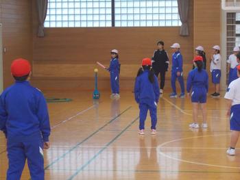 青いジャージ姿の女の子が目の前に準備されたボールをバットのようなもので打とうとし、周りでみんなが見守っている写真