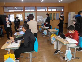 教室でマスクを着用した児童たちが机に向かって勉強しており、大人たちがその様子を見守っている写真