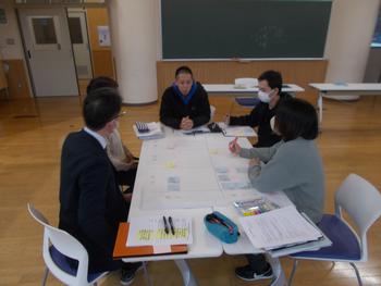 教室の一角で大人たちが円になって座り、資料を見ながら話し合いをしている様子の写真