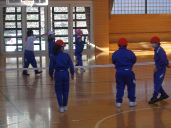 青いジャージを着て、赤い帽子をかぶった子供たちが体育館の中でバレーボールのネットの前に立っている写真