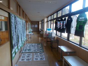 廊下の左側には、習字作品がずらりと並んで展示され、右側には、法被（はっぴ）やユニフォームのような衣装がハンガーで吊られている学校の廊下を写した写真