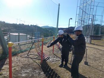 白いヘルメットと作業服を着た2人の男性が、土のグラウンドに設置された金属製の遊具を点検している様子の写真