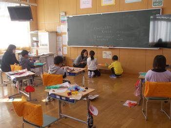 教室の前方には黒板があり、黒板の下で児童2名と先生がしゃがみこんでおり他の児童は机で作業をしている写真
