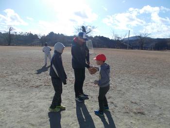 大人がグローブの使い方を指導し、他の子どもたちも順番を待っている様子の写真