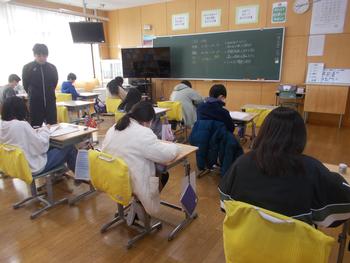 小学校の教室で窓からの自然光が差し込む中、児童が各自の机に向かって配布されたテストに回答を記入しており、教師が教室内を巡回している様子を写した写真