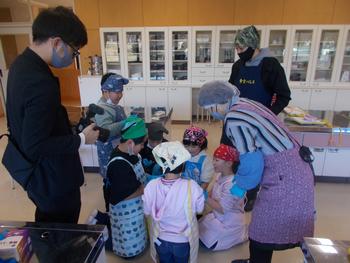 児童たちがエプロンと帽子を身につけて、円になって床に座り、数人の大人とともに室内で味噌の味見をしている写真