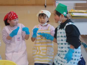 三角巾とエプロンを身に着け、青いビニール手袋をつけてた3人の児童が床に座り、みその味見をしている写真