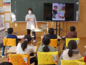 マスクをした先生が黒板の前に立ち、隣に置いたモニターに貼りだしたイラストを見せながら、席に着いた生徒たちに読み聞かせをしている様子の写真