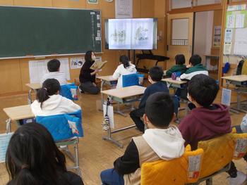 教室内で児童たちが黒板横に置いたモニターに貼りだした資料を熱心に見つめ、モニターの下にしゃがんだ先生が読み聞かせをしている授業風景の写真