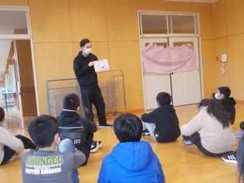 教室の床に座った生徒たちが、先生が手に持った本を見ながら話を聞いている様子の写真