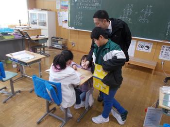 1人の女子児童が座る席に、男女の児童2人と男性の教師が集まり、先生が指差しながら説明をしている授業風景の写真