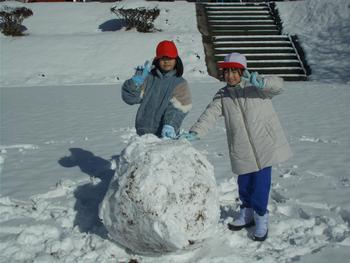 雪が一面に積もった広場で、大きな雪の玉の後ろに立った2人の女子児童が、カメラに向かって笑顔でピースサインをしている写真