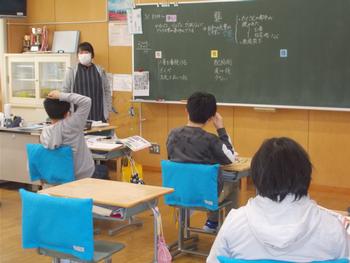 教員が黒板の前に立ち児童たちは椅子に座って前を見て授業を受けている教室内の写真