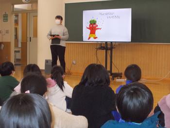 スクリーンに鬼のイラストを映し出し、先生が前に立ち、生徒たちが床に座って授業を受けている様子の写真