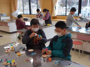 白いマスクをつけた複数の生徒たちが、テーブルの上で各自の繭細工作品を制作している授業風景の写真