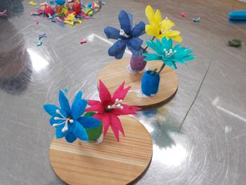 小学生が繭染めした繭を使って作った、カラフルな花瓶と花々が木製の台座の上に並べられた繭細工作品の写真