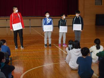 体育館の舞台前で、先生と生徒3人が肩に白い名札をかけて立ち、床に座っている他の生徒たちに向けて何かを発表している写真