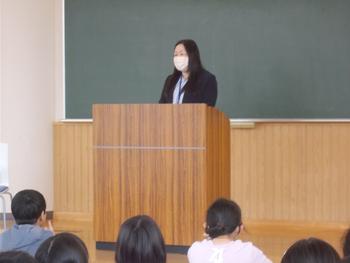 壇上に立つマスク姿の先生が、たくさんの生徒を前に話をしている様子の写真