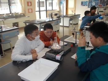 理科室で数人の小学生が実験台を囲み、先生や友達と協力しながらガラス器具を使い実験を行っている授業風景の写真
