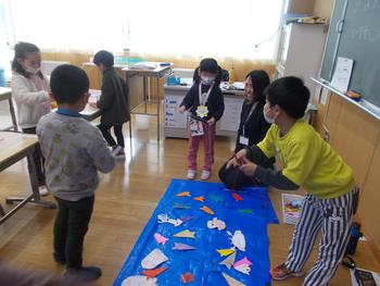 教室の床に敷かれた青いビニールシートの上で、複数の生徒が紙や画用紙で作った海の生き物を並べている授業風景の写真