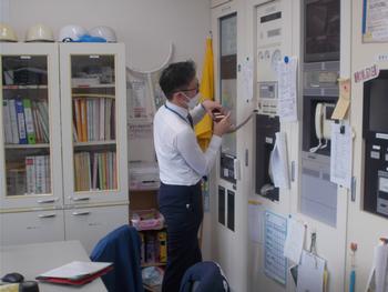 白シャツとマスク姿の男性が、オフィス内で機械の操作盤を真剣な表情で点検している様子の写真