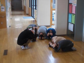 校舎の廊下で子供たちが頭を手で覆い、身を低くしてしゃがんでいる避難訓練の様子の写真