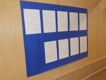 学校の廊下に設置された青い掲示板に、児童が書いたと思われる複数の手書きの文章が貼り出されている写真