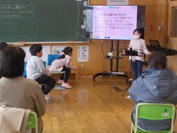 黒板とモニターがある教室で、生徒がモニターを使って発表し、他の生徒がその話を聞いている授業風景の写真