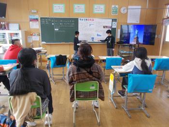 教室内で2人の生徒が黒板を使って発表し、他の生徒や先生がそれを真剣に聞いている授業風景の写真