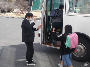 校庭に停車しているバスに乗ろうとしている小学生を先生がハイタッチで見送りしている、下校風景の写真