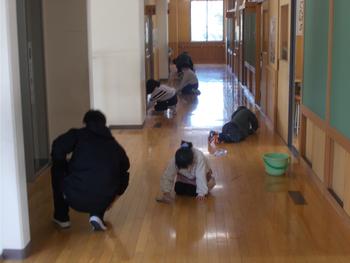 学校の廊下で複数の生徒たちが雑巾やタオルを使い、床を一生懸命に拭いている清掃活動の様子の写真