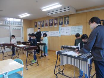 小学校の音楽室で、複数の生徒が卓上木琴を使い、先生の指導のもと真剣に演奏練習に取り組んでいる授業風景の写真