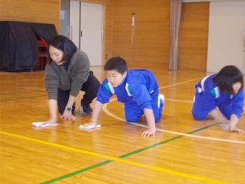 体育館の床で、先生と二人の児童が雑巾を持って四つん這いになり、床の雑巾がけをしている様子の写真