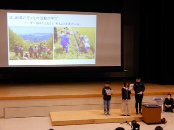 学校の体育館ステージ上で、3名の子どもたちが地域活動に関するスライドを使いながら大勢の聴衆の前で発表している様子の写真