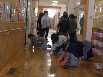 木製の床が張られた学校の廊下で、複数の小学生と先生がかがみ込んで床を拭いている様子の写真