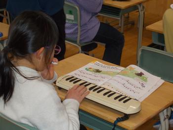 楽譜付きの教科書を見ながら女の子が鍵盤ハーモニカを吹いて演奏している様子を斜め後方から写した写真