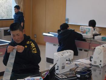 前方では男の子が布を手に取り、奥では別の児童がミシンを操作している家庭科の授業の様子を写した写真