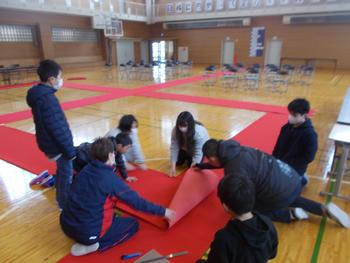 体育館で、複数の児童と先生が赤い布を床に敷き、式典の準備をしている様子の写真