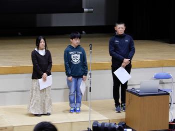 マイクの後ろに男の子と女の子の3名が1列に並んで緊張したおもむきで立っている写真