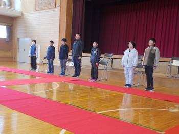 体育館の床に敷かれた赤い布の手前に置いた椅子の前で、7人の男女の児童たちが姿勢を正して立っている写真